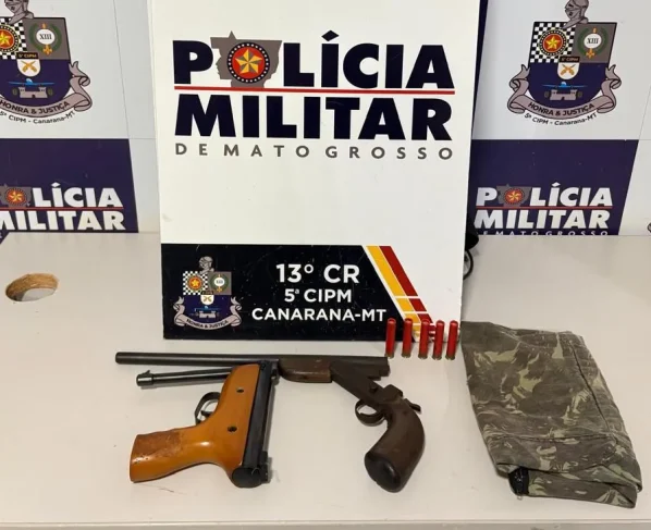 Foto das armas apreendidas em Canarana, as armas estão organizadas em uma mesa em frente à um banner com a logo da Polícia Militar de Mato Grosso