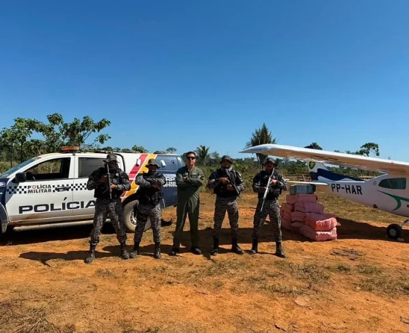 Policiais militares e da GEFRON em frente de uma viatura e avião, uma pilha de drogas apreendidas está diretamente ao lado dos agentes