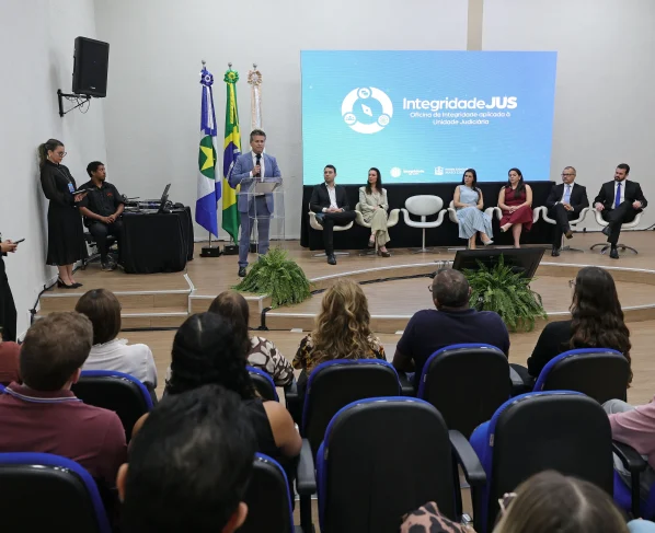 Em um auditório, várias pessoas estão sentadas em cadeiras azuis em fileiras escalonadas. Elas assistem a uma apresentação, e em primeiro plano, uma tela exibe a logo "integridade do TJMT".