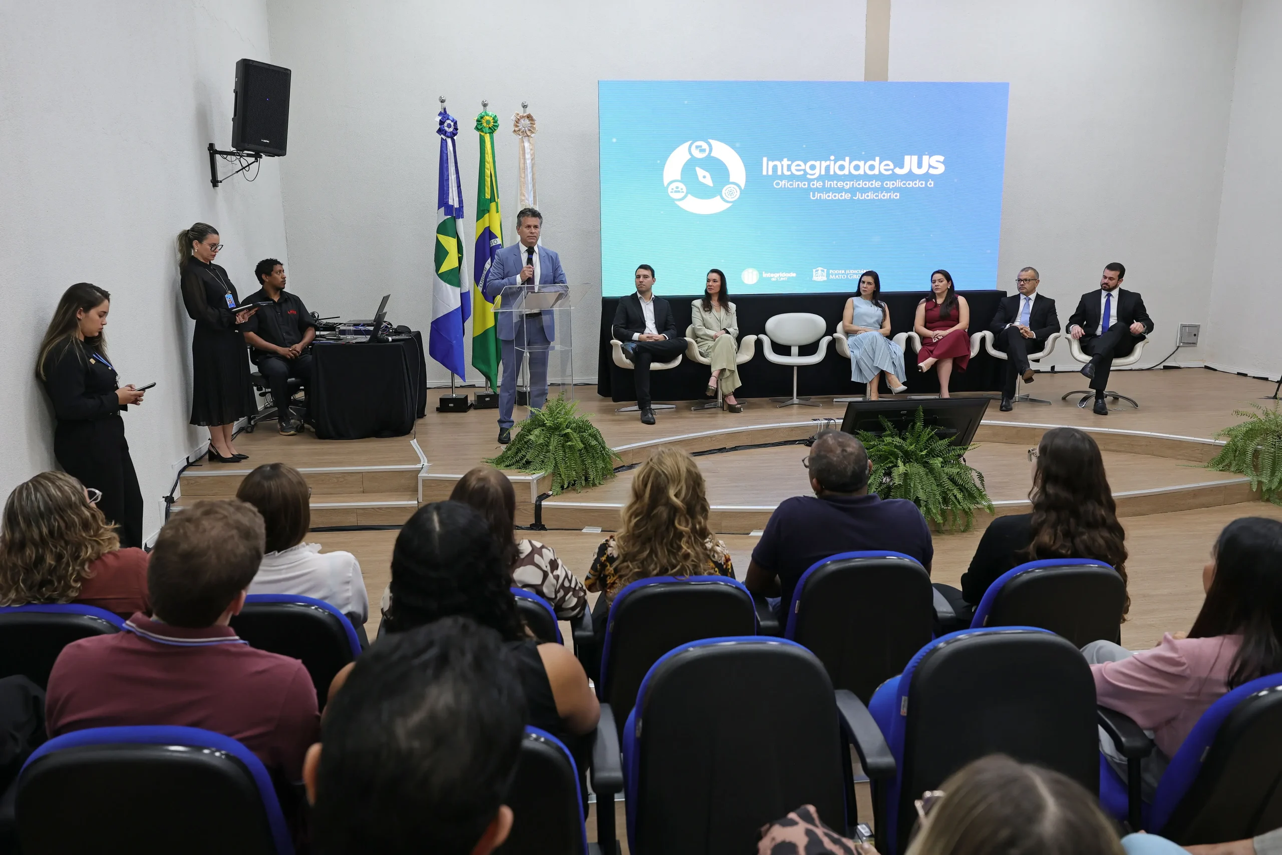 Em um auditório, várias pessoas estão sentadas em cadeiras azuis em fileiras escalonadas. Elas assistem a uma apresentação, e em primeiro plano, uma tela exibe a logo "integridade do TJMT".