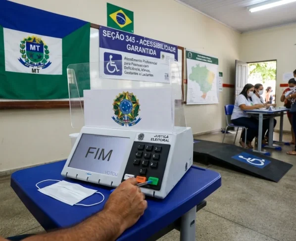 Urna eletrônica em seção eleitoral com sinalização de acessibilidade e bandeira institucional